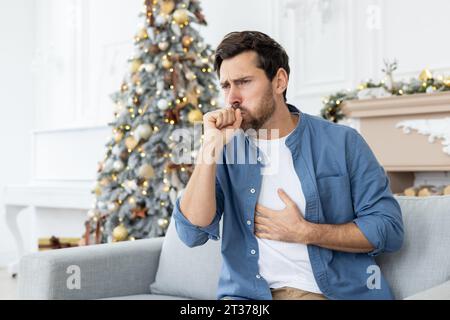 Porträt eines jungen Mannes, der während der festlichen Weihnachtsfeiertage und des Wochenendes erkrankt ist. Zu Hause auf dem Sofa sitzen und Husten, seinen Mund mit der Hand bedecken und seine Brust halten. Stockfoto