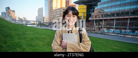 Glückliches asiatisches Mädchen steht auf der Straße, Universitätsstudentin läuft mit einem digitalen Tablet in den Händen und lächelt, steht im Stadtzentrum Stockfoto