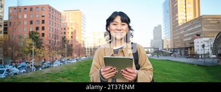 Glückliches asiatisches Mädchen steht auf der Straße, Universitätsstudentin läuft mit einem digitalen Tablet in den Händen und lächelt, steht im Stadtzentrum Stockfoto