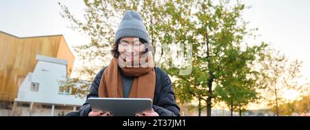 Porträt eines asiatischen Mädchens in warmer Kleidung, sitzt auf einer Bank mit digitalem Tablet und Stift, lächelt fröhlich und zieht bei kaltem Wetter ins Freie Stockfoto