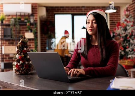 Asiatische Angestellte mit weihnachtsmann-Hut, die im saisonalen Festbüro im Winter an einem Laptop arbeitet. Firmenleiter sitzt am Schreibtisch und benutzt am weihnachtlich dekorierten Arbeitsplatz den Computer Stockfoto