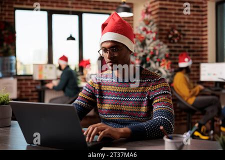 Junger afroamerikanischer Mann mit weihnachtsmütze, der die Arbeit auf dem Laptop im weihnachtlich dekorierten Büro verwaltet. Mitarbeiter des Unternehmens, der während der Winterferien am Arbeitsplatz arbeitet Stockfoto