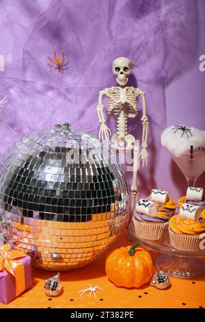 Halloween Party Komposition mit Cupcakes, Discokugel und Dekorationen auf orangem Tisch vor violettem Hintergrund Stockfoto