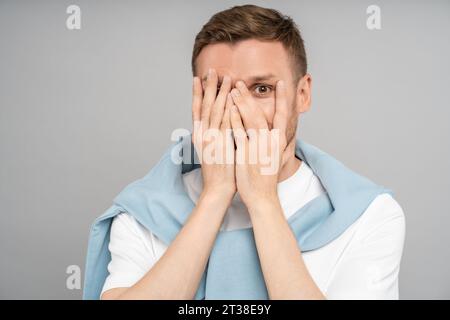 Schüchterner bescheidener verspielter Kerl, der das schließende Gesicht mit den Händen versteckt, ein Auge, das auf grauen Hintergrund in die Kamera schaut. Stockfoto