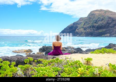 Eine junge Frau im lila Kleid sitzt am felsigen Ufer und blickt auf den Ozean am Makapu'u Beach in Oahu, Hawaii Stockfoto
