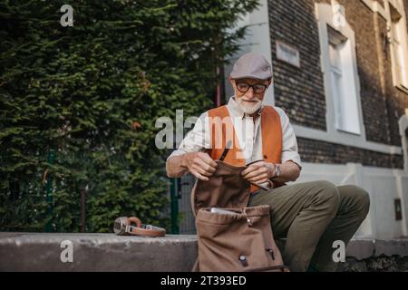 Senior-Mann sitzt auf Straßenrand und arbeitet an Laptops mit digitalen Technologien. Seniorenkonzept und digitale Kompetenzen. Stockfoto