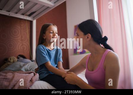 Mutter weckt die Tochter morgens zur Schule auf. Sie kniete am Kinderbett und hielt die Tochter mit den Händen. Stockfoto