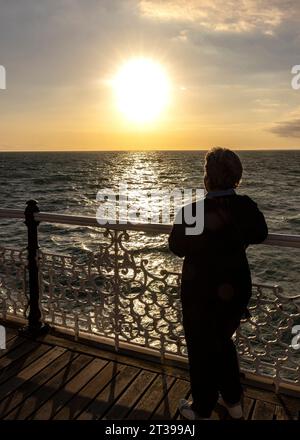 Eine Dame, die nachdenklich, entspannend und die untergehende Sonne über dem Meer vom Brighton Palace Pier aus genießt Stockfoto