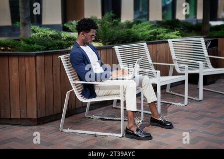 Junger afroamerikanischer Geschäftsmann mit Laptop, während er auf der Bank in der Stadtstraße sitzt. Lebensstil beruflicher männlicher Arbeit im städtischen Umfeld. Stockfoto