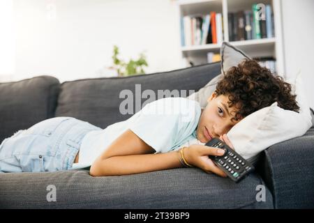 Ein zehnjähriges langweiliges Mädchen, das auf einem Sofa liegt und fernsehen sieht, mit einem tv-Controller, in einem Wohnzimmer. Ein Mädchen, das zu Hause einen Film ansieht. Stockfoto