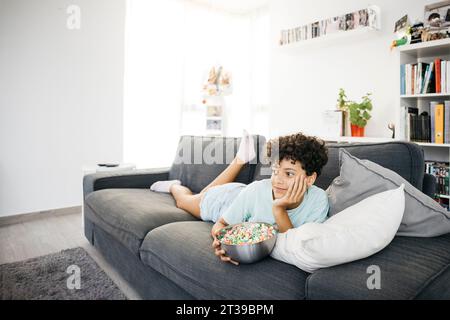 Ein zehnjähriges langweiliges Mädchen, das auf einem Sofa liegt und fernsehen sieht, mit einem tv-Controller, in einem Wohnzimmer. Ein Mädchen, das zu Hause einen Film ansieht. Stockfoto