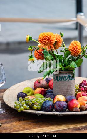 Leuchtend orange Dahlien in einem rustikalen Topf auf einem Holztisch neben einer Platte mit frischem Obst. Stockfoto
