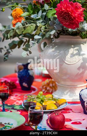Eine farbenfrohe Tischdekoration mit Weingläsern, Früchten auf einem Teller und einer blau verzierten Vase vor einem rot gemusterten Tuch. Stockfoto