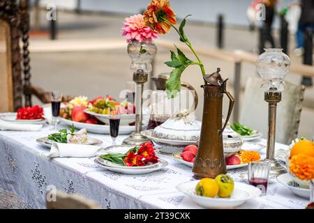 Ein eleganter Esstisch im Freien mit frischem Obst, verzierten Gerichten, Vintage-Laternen, einem Messingkrug und einer lebendigen Dahlienblume auf einem Spitzenetikett Stockfoto