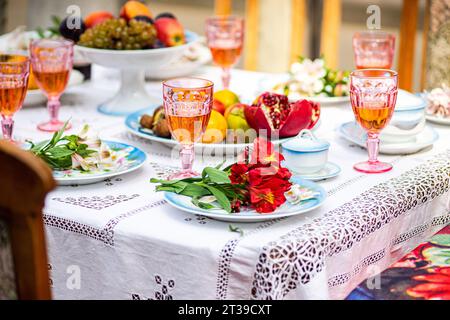 Der Esstisch in hellen Farben ist mit rosafarbenen Getränken in Kristallgläsern eingerichtet, ein Teller mit roten Blumen, verschiedenen Früchten und kunstvollen Porzellan-Desserts Stockfoto