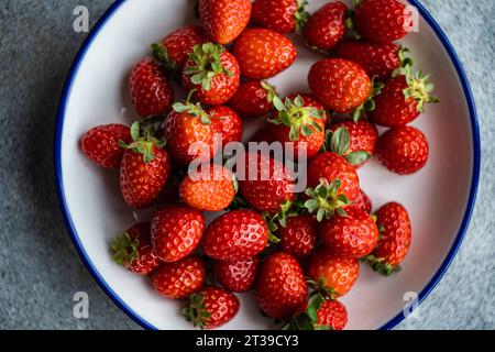 Blick von oben auf leuchtend rote Bio-Erdbeeren mit grünen Blättern, auf einer weißen Platte mit blauem Rand, vor einem strukturierten grauen Hintergrund. Stockfoto