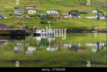 Malerischer Blick auf kleine Dorfhäuser an der grünen Küste des ruhigen Sees mit schwimmender Yacht auf Färöern Stockfoto