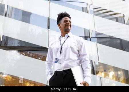 Niedriger Winkel eines selbstbewussten afroamerikanischen männlichen Unternehmers in formeller Kleidung in der Nähe eines modernen Gebäudes mit Hand in der Tasche, während er den Laptop hält Stockfoto