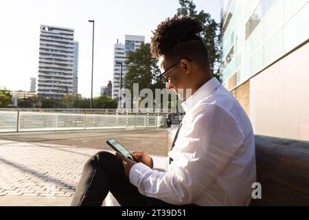 Seitenansicht eines afroamerikanischen männlichen Unternehmers in formeller Kleidung und Brille, der auf der Bank sitzt und auf dem Smartphone surft, während er auf der Straße der Stadt ruht Stockfoto