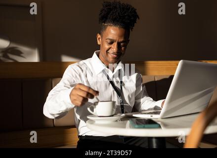 Glücklicher junger afroamerikanischer Freelancer in lässigen Klamotten und rasierten Dreadlocks, der am Kaffeetisch mit einer Tasse Kaffee sitzt, während er im Laptop surft Stockfoto