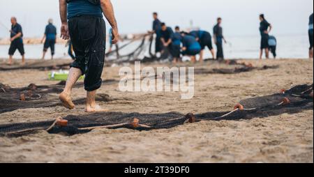 Rückansicht eines anonymen Mannes in nasser Kleidung, während er am Sandstrand ein Fischernetz gegen verschwommene Fischer mit Fang aufstellt Stockfoto