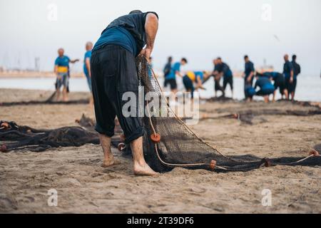 Rückansicht eines anonymen männlichen Mannes in nasser Kleidung, der nach unten blickt, während er am Sandstrand ein trocknendes Fischernetz gegen verschwommene gesammelte Fischer mit Fang hält Stockfoto