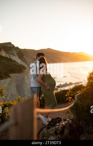 Seitenansicht einer anonymen schwangeren Frau, die am Abend einen unerkennbaren Mann küsst, während sie an der Küste in Seehöhe steht Stockfoto