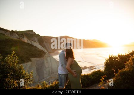 Seitenansicht einer anonymen schwangeren Frau, die am Abend einen unerkennbaren Mann küsst, während sie an der Küste in Seehöhe steht Stockfoto