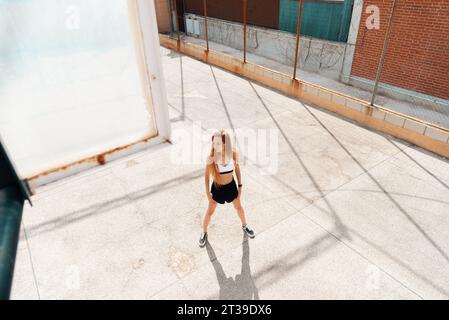 Junge rothaarige Frau, die sich an einem sonnigen Tag im Freien ausdehnt, einen weißen Sport-BH und schwarze Shorts trägt, mit einem Straßenbelag Hintergrund. Stockfoto