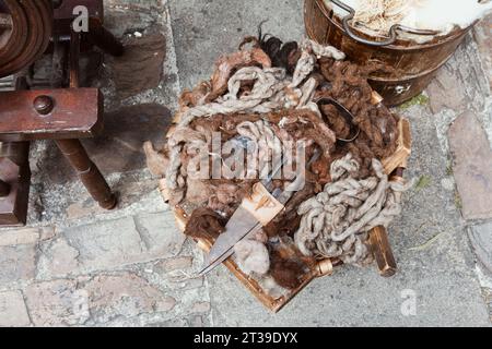 Nahaufnahme eines Vintage-Spinnrads und Werkzeuge umgeben von braunen Wollklumpen auf einem rustikalen Steinboden. Stockfoto