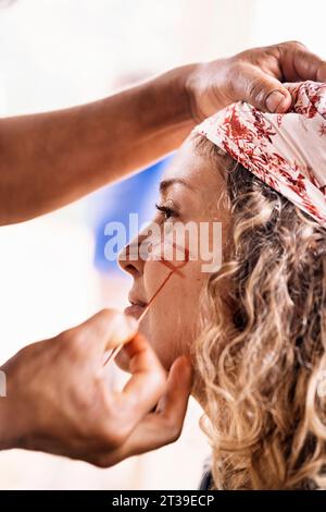 Ernten Sie anonyme Person, die traditionelle Stammesfarbe auf das Gesicht einer lächelnden blonden Frau mittleren Alters vor verschwommenem Hintergrund in der Amazonasstraße aufträgt Stockfoto