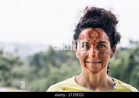 Porträt einer lächelnden Frau mittleren Alters mit Farbe im Gesicht, während sie legere Kleidung trägt, vor verschwommenem grünem Hintergrund steht und in die Kamera blickt Stockfoto