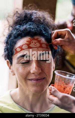 Ernten Sie anonyme Person, die traditionelle Stammesfarbe auf die Stirn einer lächelnden Frau mittleren Alters mit geschlossenen Augen vor verschwommenem Hintergrund in Amazo aufträgt Stockfoto