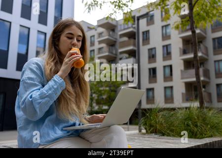 Junge blonde Studentin trinkt Orangensaft und arbeitet in ihrem Laptop in der Stadt. Stockfoto