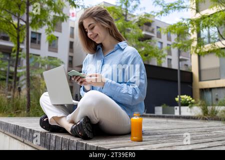 Eine junge, glückliche blonde Studentin sitzt auf einer Holzbank auf einem Stadtplatz und benutzt gleichzeitig ihr Handy und ihren Laptop. Stockfoto