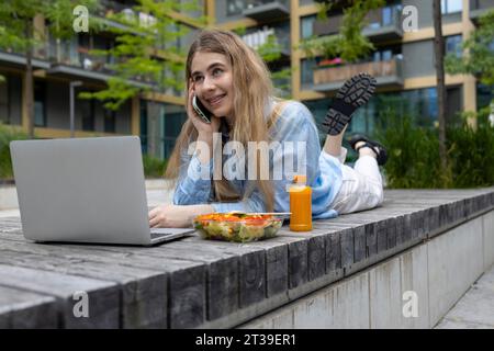 Glückliche blonde Studentin mit Zahnspangen, die in einer Holzbank liegt und mittags in der Stadt mit ihrem Smartphone spricht. Stockfoto