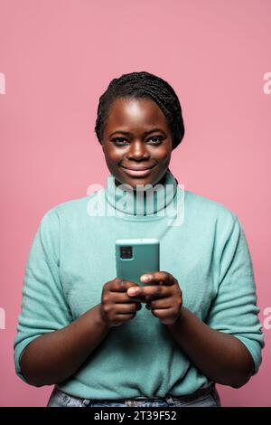 Eine fröhliche Afrikanerin in einem blauen Pullover, die ein Smartphone mit rosa Hintergrund hält Stockfoto