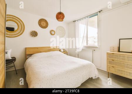 Komfortables Bett mit Holzkommode und Dekorationen an weißen Wänden in stilvollem Schlafzimmer mit Fenster und weißen Wänden Stockfoto