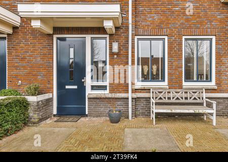 Äußere des Backsteinhauses mit blauer Eingangstür und Fenstern und leerer Bank auf der gepflasterten Terrasse Stockfoto