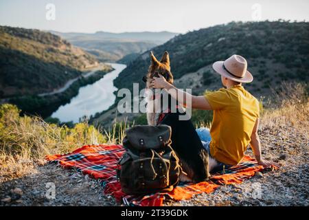 Rückansicht eines anonymen männlichen Wanderers in gelblichem Hemd, der auf einem grünen Hügel mit einem bezaubernden Hund in rotem Kragen sitzt und die malerische Landschaft bewundert Stockfoto