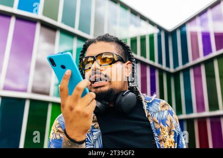 Lateinischer Mann mit Zöpfen, der mit seinem blauen Smartphone vor einem bunten Gebäude in der Stadt mit Sonnenbrille und Kopfhörern ein Gespräch führt. Stockfoto
