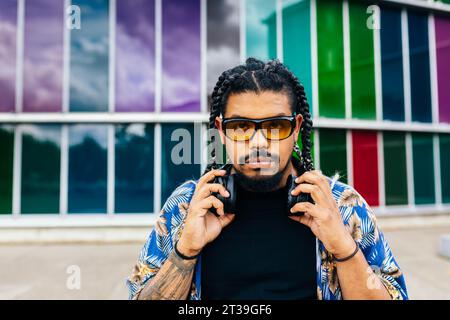 Lateinischer Mann mit Zöpfen und Sonnenbrille, der Kopfhörer um den Hals hält und vor einem mehrfarbigen Hintergrund auf die Vorderseite blickt. Stockfoto