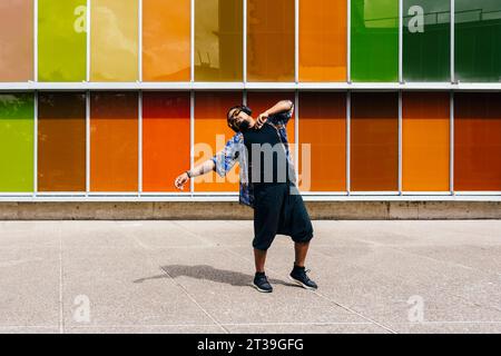 Ein lateinischer Mann in Sonnenbrille und Kopfhörern, der vor einem städtischen Gebäude aus bunten Paneelen tanzt. Stockfoto