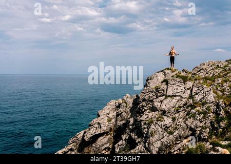 Ein fitter Mann spielt eine Yoga-Pose auf einer felsigen Klippe mit Blick auf das Meer. Stockfoto