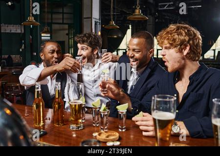 Aufgeregte multiethnische Freunde halten Tequila mit Limette neben Gläsern Bier auf der Bar Stockfoto