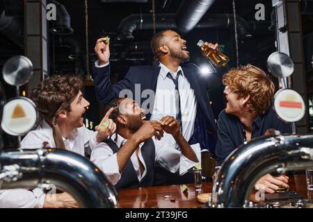 Glückliche Männer, die einen afroamerikanischen Freund sehen, der Bier aus zwei Flaschen trinkt, nachdem er in der Bar gearbeitet hat Stockfoto