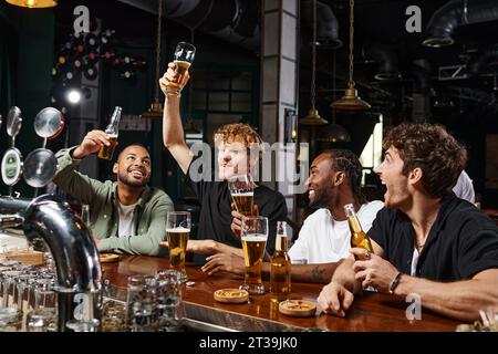 Gruppe von vier glücklichen, multiethnischen männlichen Freunden, die an der Bar ein Glas Bier heben, Junggesellenabschied Stockfoto