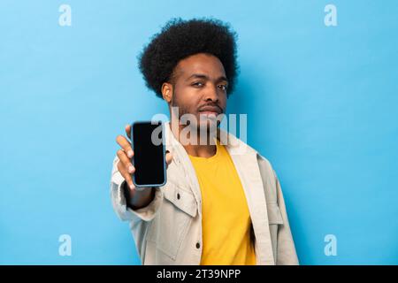 Afro-amerikanischer Mann, der ein Mobiltelefon mit leerem Bildschirm in isoliertem hellblauem Farb-Studio-Hintergrund zeigt Stockfoto