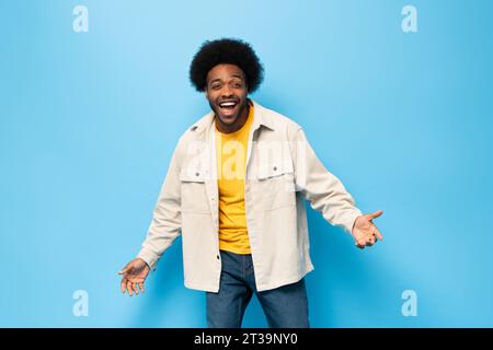 Überraschter afro-amerikanischer Mann lächelt mit geöffneter Hand Geste in hellblauem Farbstudio fotografiert isolierten Hintergrund Stockfoto