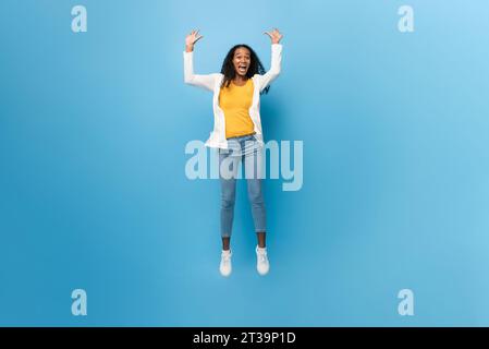 Porträt der lächelnden ekstatischen afroamerikanischen Frau, die mit Armen in isoliertem hellblauem Farb-Studiohintergrund springt Stockfoto